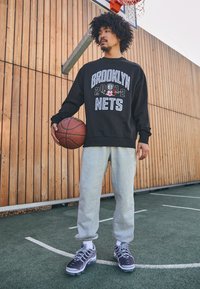 Felpa nera con grafica "BROOKLYN NETS 2012", pantaloni della tuta grigi e scarpe da ginnastica grigie, che tiene un pallone da basket, su un campo all'aperto.