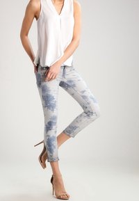 Jeans de patrón floral gris claro con una textura suave, que presentan diseños de árboles azules. Combinados con una blusa sin mangas blanca y tacones beige.