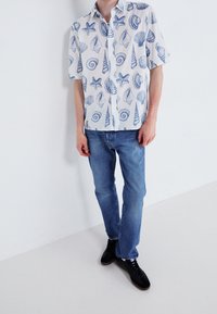 Košile s krátkým rukávem má bílý základ s modrými vzory mořských lastur a hvězdic; doplněná džínami a černými teniskami.