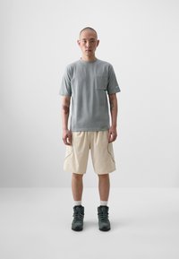 T-shirt gris à col rond avec une poche poitrine, associé à un short beige clair doté de coutures latérales contrastées. Des bottes sombres complètent le look.