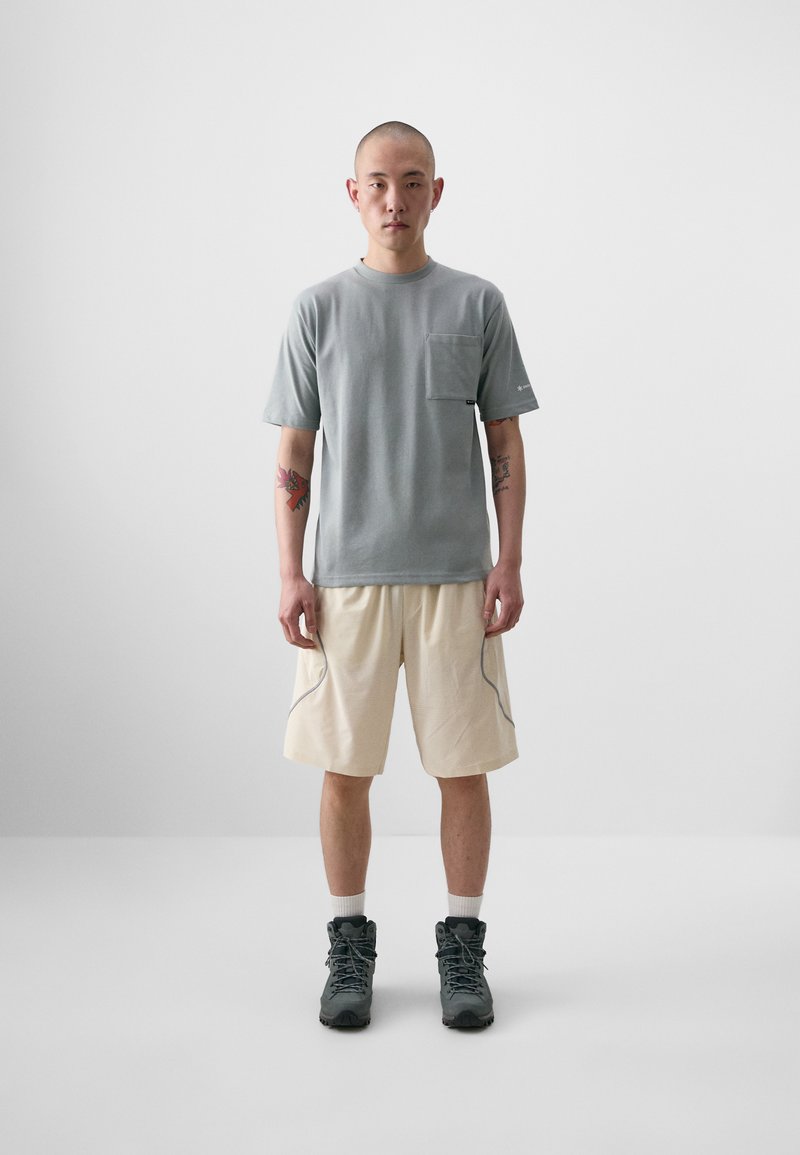 T-shirt gris à col rond avec une poche poitrine, associé à un short beige clair doté de coutures latérales contrastées. Des bottes sombres complètent le look.