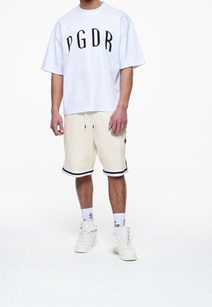 T-shirt blanc oversize avec texte imprimé noir "PGDR." Shorts crème avec bandes latérales noires. Baskets blanches et chaussettes avec texte noir.