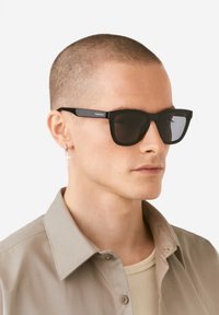 Black square sunglasses with dark lenses and thick plastic frames. Branded detail on the arm. Worn by a person with short hair and a beige shirt.