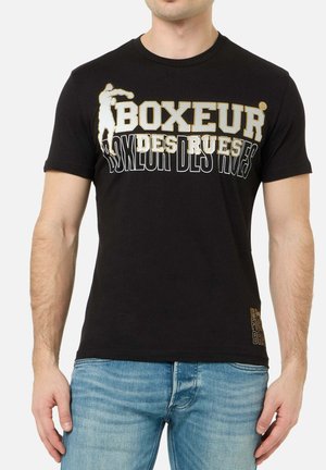 Mann trägt schwarzes T-Shirt mit gold-weißem "Boxeur Des Rues" Schriftzug und einer Boxer-Grafik, kombiniert mit blauer Jeans.