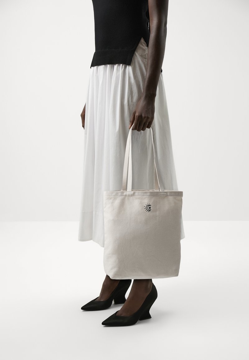 Personne en haut noir sans manches et jupe blanche tient un sac cabas beige avec un petit symbole de soleil et de lune, portant des talons noirs.