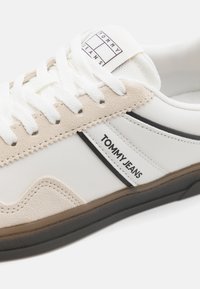 Fehér sneaker bézs velúr kiegészítőkkel. Fekete csíkkal és "TOMMY JEANS" logóval rendelkezik. Textúrált talp és pamut fűző. Lazább dizájn.