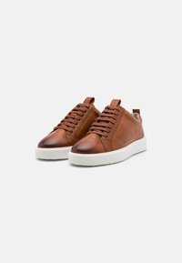 Brune lærersneakers med en glat overflade, flade hvide gummisåler, runde tæer og teksturerede snørebånd. Har subtile perforeringer på siderne.