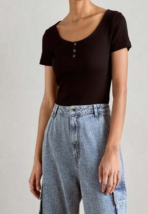 Femme portant un haut noir à manches courtes avec des détails de boutons et un pantalon large bleu clair texturé taille haute avec des poches.
