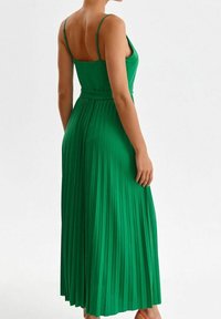 Grön plisserad maxiklänning med tunna axelband, cinchad midja och flödande kjol. Tyget ser slätt ut med vertikala veck som förstärker texturen.