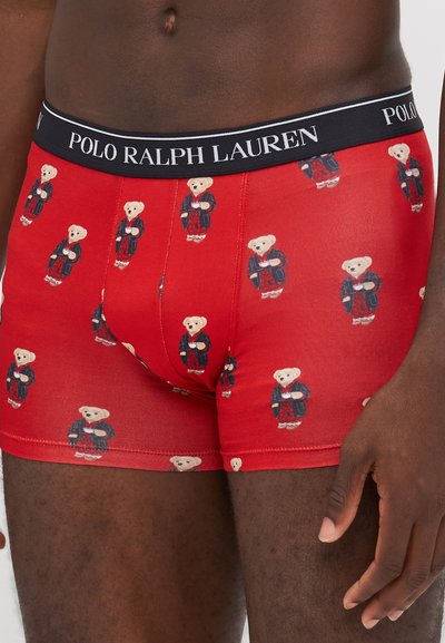 Sarkanas bokseršortas ar atkārtotu teddy lāča rakstu, kas ģērbti tumši zilos tērpos, ar melnu jostu, uz kuras izšūts "POLO RALPH LAUREN" logs.