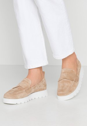 Beige Wildleder-Slipper mit einer strukturierten Oberseite und einer dicken weißen Gummisohle, mit einer dekorativen Naht und einer glatten Oberfläche.