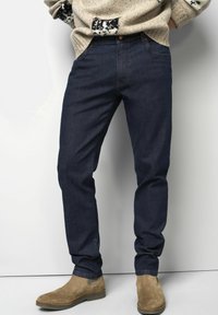 Jean en denim bleu foncé à coupe slim, doté de cinq poches et d'une braguette zippée. Associé à des bottines en suède marron clair.