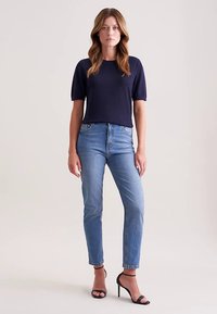 Granatowy top z krótkim rękawem w połączeniu z wysokim stanem niebieskich dżinsów i czarnymi sandałkami na obcasie, stojąca na neutralnym tle.