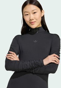 Svart långärmad topp med hög hals, volangärmar och en diskret Adidas-logga på bröstet. Tillverkad av ett mjukt, elastiskt material.