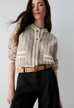 Frau trägt eine beige strukturierte, geknöpfte Strickjacke mit Taschen, einen braunen Gürtel und schwarze, hoch taillierte Hosen vor einem schlichten hellen Hintergrund.