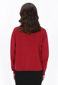 Pullover tricotat roșu cu un croi lejer, mâneci lungi și manșete cu nervuri. Textură netedă, lungime scurtă și detalii discrete ale cusăturilor la spate.
