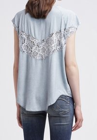 Světle modrý top s krátkým rukávem a krajkovým vložením ve vzoru šípu na zadní straně. Hladký materiál s mírně zakřiveným spodním lemem.