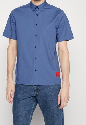 Mand iført blå kortærmet skjorte med sorte knapper og rød Hugo-logo badge, kombineret med blå jeans.