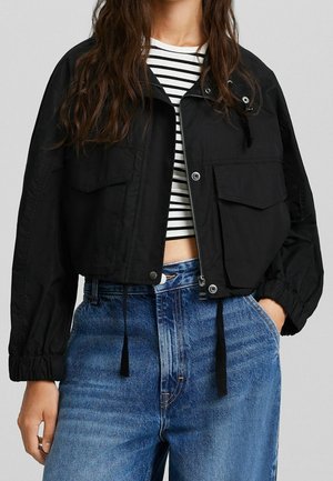 Frau trägt eine kurze schwarze Jacke mit großen Fronttaschen über einem schwarz-weiß gestreiften kurzen Top und hoch taillierten blauen Jeans.