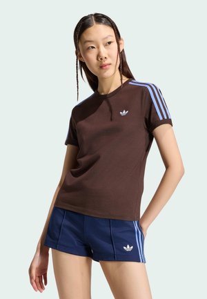 Joven mujer con el cabello trenzado lleva una camiseta marrón de Adidas con rayas azul claro y pantalones cortos azul marino con rayas y logo a juego.