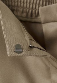 Gros plan sur la taille d'un pantalon beige avec un petit crochet métallique argenté et une fermeture éclair partiellement ouverte sur un tissu texturé.