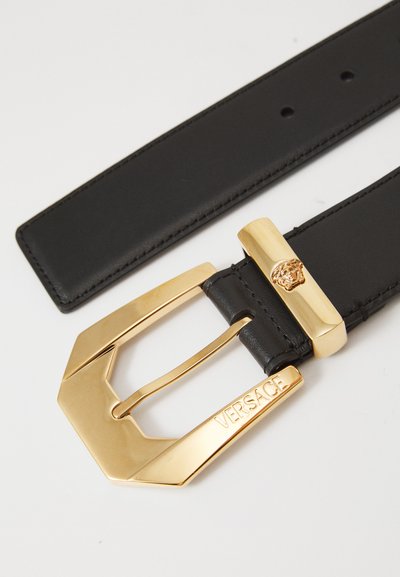 Black leather belt with a large gold geometric buckle. Buckle features a lion head accent and “VERSACE” engraved. Smooth texture throughout.