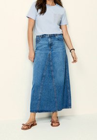 Denim-maksihame, jossa on korkea vyötärö, sininen väri, etusauman yksityiskohta, yhdistettynä vaaleansiniseen t-paitaan, jossa on raidallinen tasku. Ruskeat sandaalit.