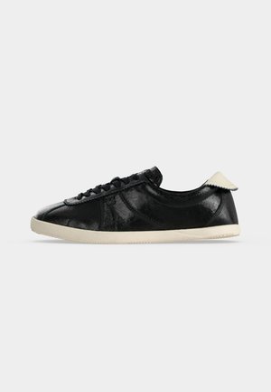 Sort lavtopet sneaker med hvid sål og hælfane, tekstureret læderoverdel og sorte snørebånd på en ensfarvet lys baggrund.