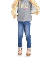 Gul, glänsande jacka över en svart och vit randig skjorta med texten "GIRLS". Blå jeans och ljusbruna sandaletter med remmar fulländar outfiten.