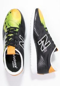 Baskets de sport noires avec un motif vibrant vert et jaune sur le bout, présentant une surface texturée, une semelle blanche et un logo "N" bien visible.