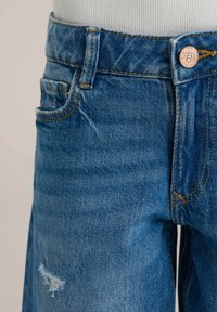 Blugi din denim albastru cu finisaj estompat, având talie înaltă, cusături aurii și un detaliu uzat pe coapsa stângă. Nasture metalic la talie.