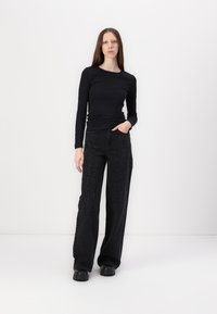 Zwarte langslijf top gecombineerd met wijde zwarte broek met studs. Model staat met één hand in de zak tegen een witte achtergrond.