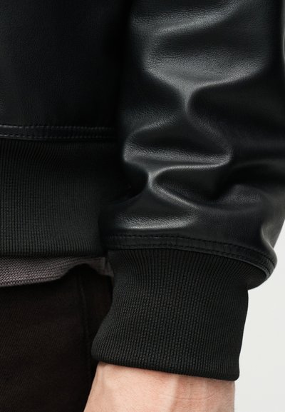 Κοντινό πλάνο στον καρπό ενός ατόμου που φοράει μαύρο δερμάτινο μπουφάν με μανσέτες από ριμπ πλεκτό και γκρι πουκάμισο που φαίνεται από κάτω.