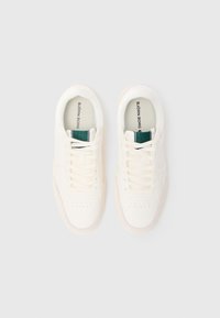 Sneakers brancos com parte superior em canvas texturizado, frente com atacadores e detalhes perfurados. Acento verde na língua. Sola macia. Design minimalista.