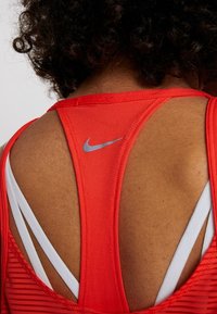 Gros plan sur le haut du dos d’une personne portant un haut de sport Nike rouge avec un logo Swoosh gris et des bretelles de soutien-gorge blanches visibles.