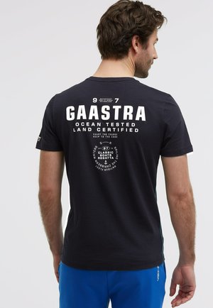 Mann von hinten, trägt ein schwarzes Gaastra-T-Shirt mit weißem nautischem Schriftzug und blaue Shorts vor einfarbig hellem Hintergrund.