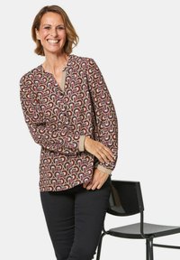 Braun gemusterte Bluse mit abstrakten Formen in Rot und Weiß; verfügt über einen V-Ausschnitt, Knopfdetails und lange Ärmel. Kombiniert mit schwarzen Hosen.