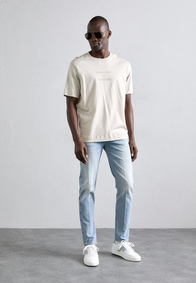 Μπεζ βαμβακερό t-shirt με ανάγλυφο λογότυπο, συνδυασμένο με ανοιχτό γαλάζιο τζιν και λευκά αθλητικά παπούτσια. Αξεσουάρ περιλαμβάνουν σκούρα γυαλιά ηλίου.