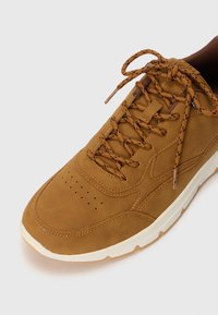 Brown suede sneaker with a rounded toe, perforations on the toe box, and patterned laces. Features a white rubber sole and subtle stitching details.