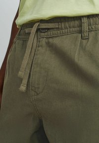 Beżowe spodnie wykonane z miękkiego materiału, z elastycznym paskiem w talii i sznurkiem do regulacji. Projekt zawiera boczne kieszenie oraz ozdobny guzik.