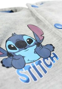 Tissu gris avec un personnage de dessin animé bleu aux grands yeux et aux oreilles roses. Le texte « STITCH » est imprimé en bleu sous le personnage.