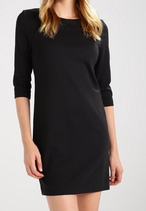 Frau trägt ein einfaches, knielanges schwarzes Kleid mit dreiviertel Ärmeln und rundem Ausschnitt, steht vor einem hellen Hintergrund.