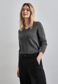 Grauer Strickpullover mit V-Ausschnitt, langen Ärmeln und Rippbündchen, kombiniert mit schwarzen Hosen, präsentiert einen lässigen Schnitt und eine glatte Textur.