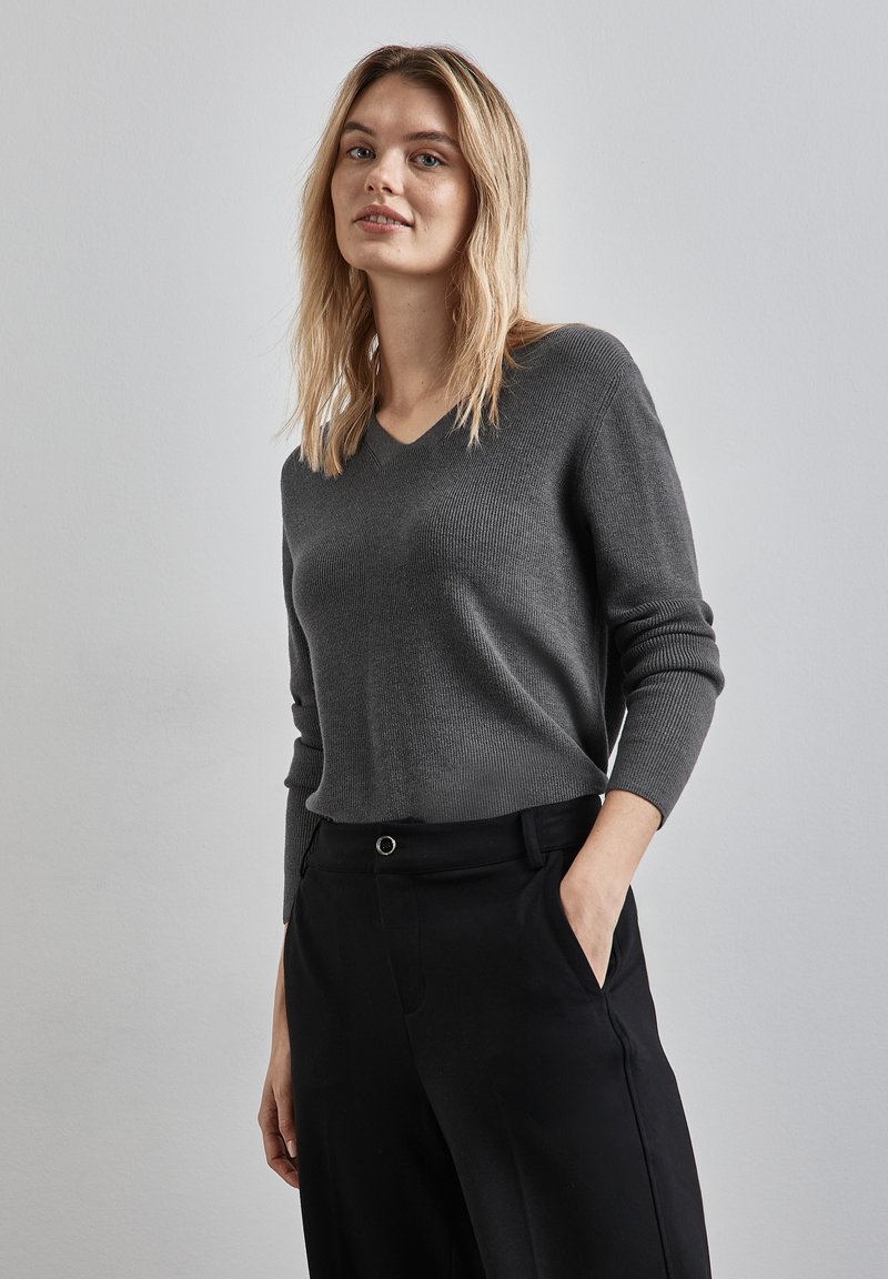 Grauer Strickpullover mit V-Ausschnitt, langen Ärmeln und Rippbündchen, kombiniert mit schwarzen Hosen, präsentiert einen lässigen Schnitt und eine glatte Textur.