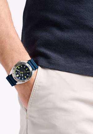 Horloge met een zilveren roestvrijstalen kast, een zwarte wijzerplaat met gele accenten en een blauwe rubberen band; gedragen om een pols met een grijs shirt.