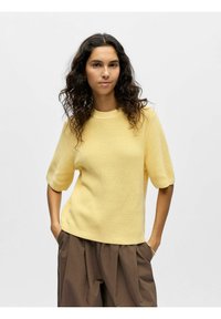 Pull en tricot jaune à col rond, manches courtes bouffantes et texture côtelée, associé à un pantalon large marron.