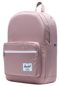 Sac à dos rose clair avec un dessus arrondi, une poche zippée à l'avant et un écusson en tissu. Texture lisse, design minimaliste, bretelles ajustables.