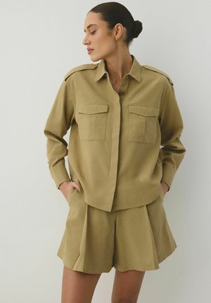 Kobieta z ciemnymi włosami w kok, ubrana w khaki koszulę zapinaną na guziki z kieszeniami na piersi oraz dopasowane plisowane szorty, ręce w kieszeniach, patrzy na bok.