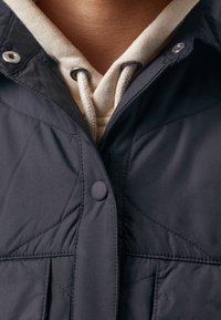 Chaqueta acolchada en azul marino oscuro con textura acolchada, cierre de botones a presión y capucha beige fija, que cuenta con cordones y detalles cosidos.