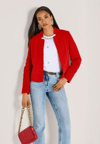 Rote strukturierte Jacke mit kurzem Schnitt, getragen über einem weißen Oberteil mit Verzierungen, kombiniert mit hellblauen Schlagjeans und einer roten Handtasche.